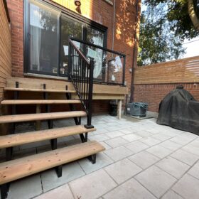 Modern wooden deck with a black glass railing and steps leading to a backyard.