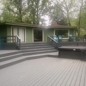 Green cabin with expansive grey deck featuring various outdoor furniture and hot tub