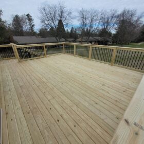 Spacious elevated wooden deck in a backyard with a fenced perimeter and lush trees in the background