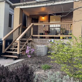 Wooden porch with stairs leading up to a cozy house entrance, surrounded by lush green bushes.