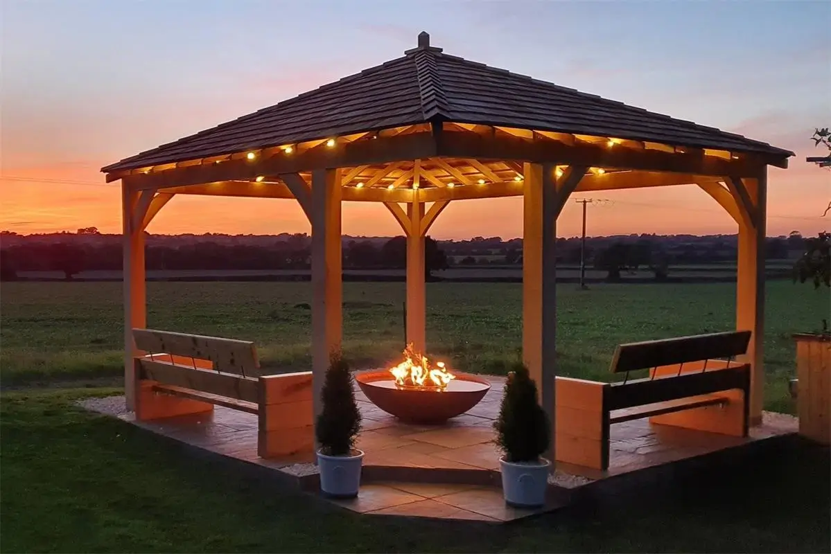 Rustic outdoor pavilion with stone fireplace, wooden beams, bar seating, and ambient lighting in a landscaped backyard