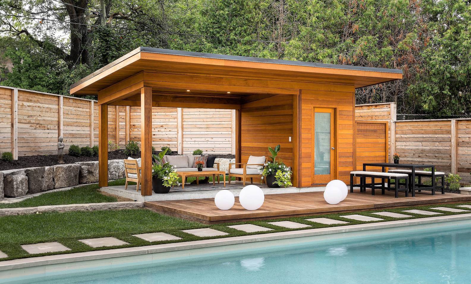 Modern cabana with lounge seating and curtains beside illuminated swimming pool at night.
