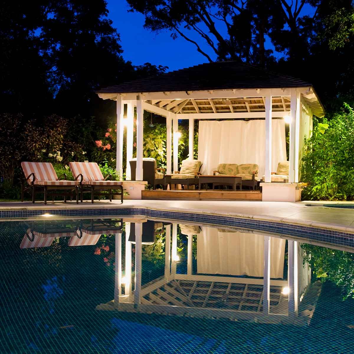 Modern cabana with lounge seating and curtains beside illuminated swimming pool at night.