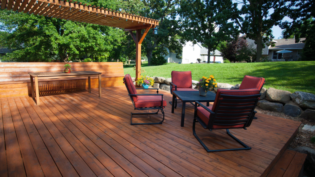 Wooden backyard deck with pergola, red cushioned patio chairs, outdoor table, and vibrant flower planters"