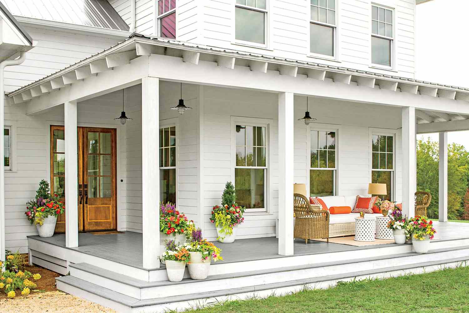 Charming farmhouse porch with white siding, wooden front door, potted flowers, and wicker seating.