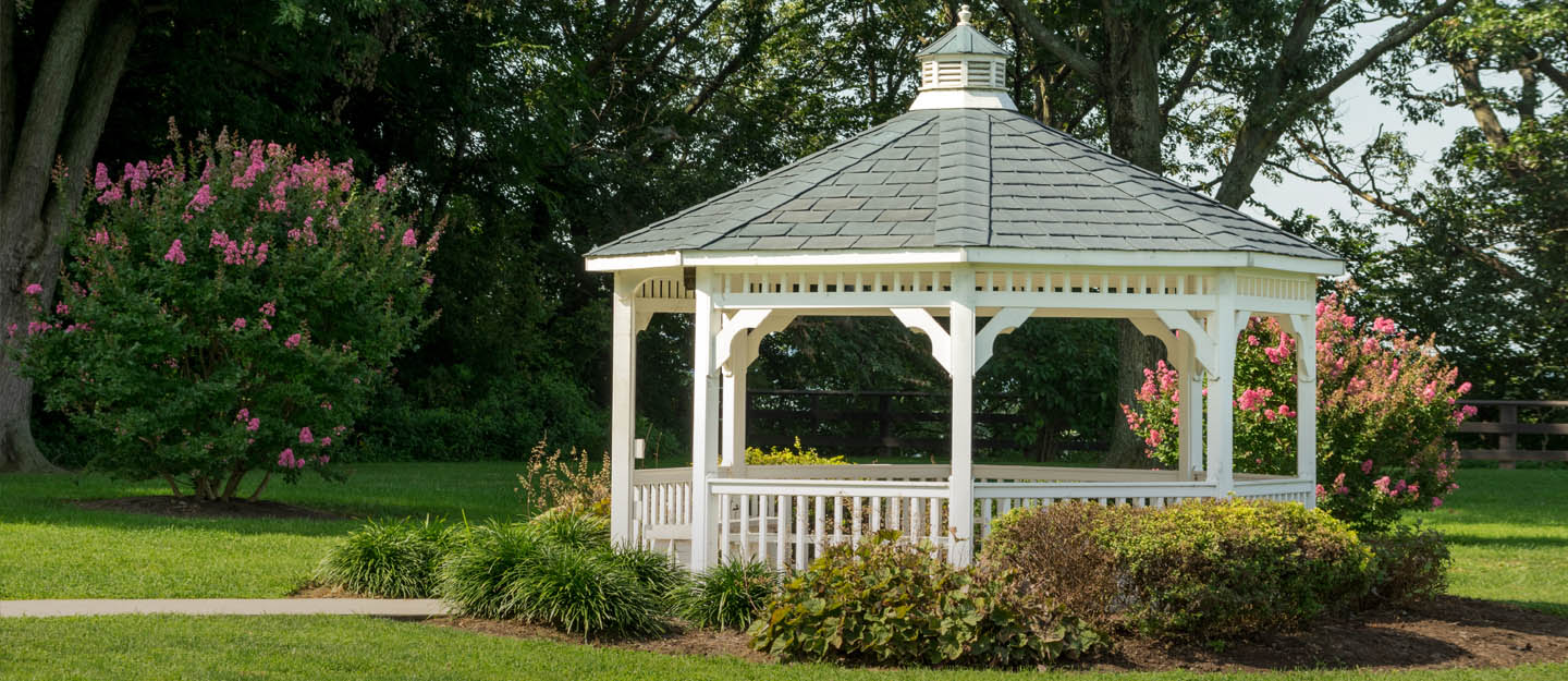 Classic white gazebo with shingled roof in a landscaped garden surrounded by greenery and flowers.