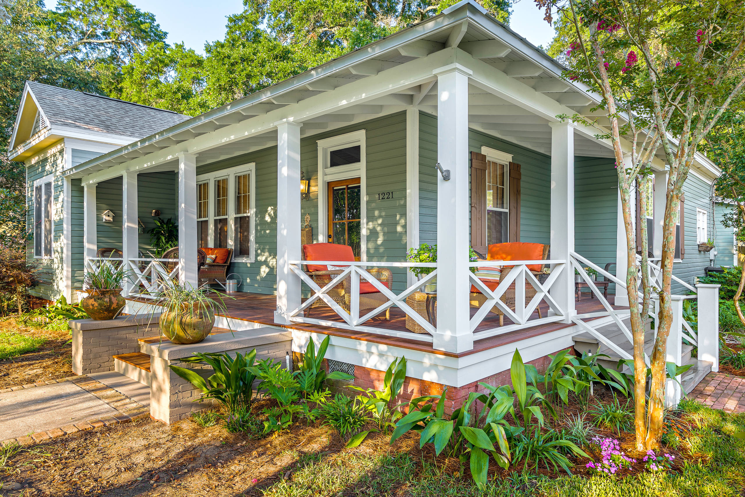 Inviting cottage-style porch with green siding, white trim, potted plants, and wicker chairs with orange cushions.