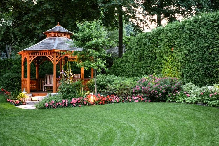 Gazebo with shingled roof in a landscaped garden surrounded by greenery and flowers.