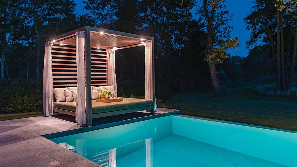 Modern cabana with lounge seating and curtains beside illuminated swimming pool at night.