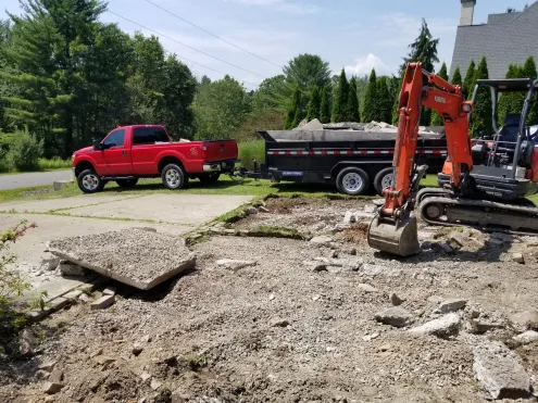 Selective demolition of concrete driveway using compact excavator with dump trailer and truck nearby.