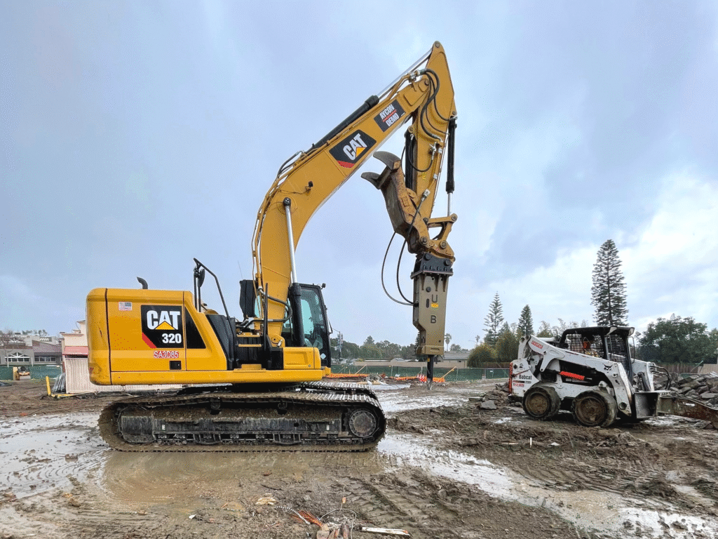 Excavator with hydraulic breaker and skid steer loader at a commercial demolition site.