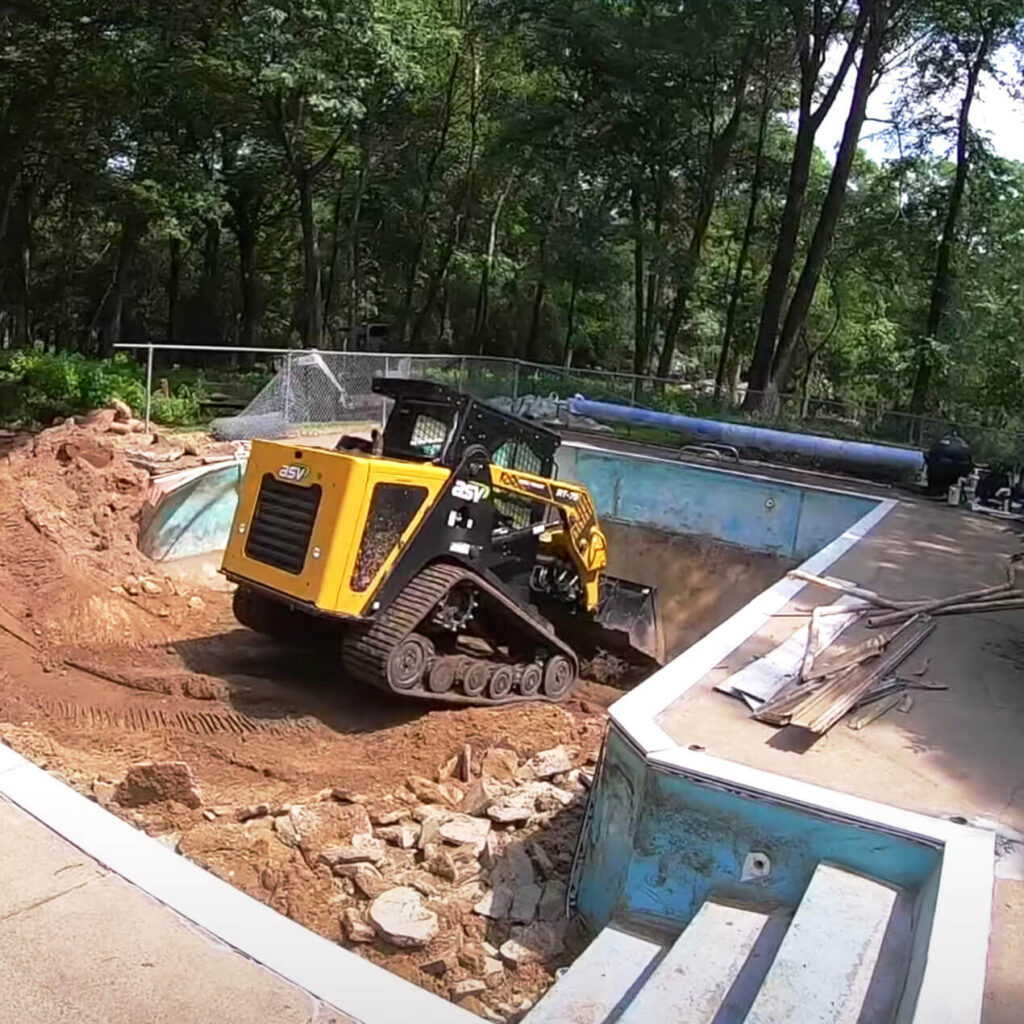 Selective demolition of concrete driveway using compact excavator with dump trailer and truck nearby.
