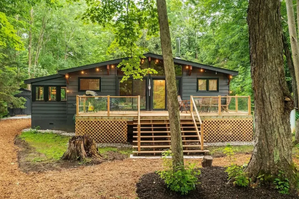 Modern black cabin-style house with wooden deck and outdoor seating, surrounded by lush green forest.