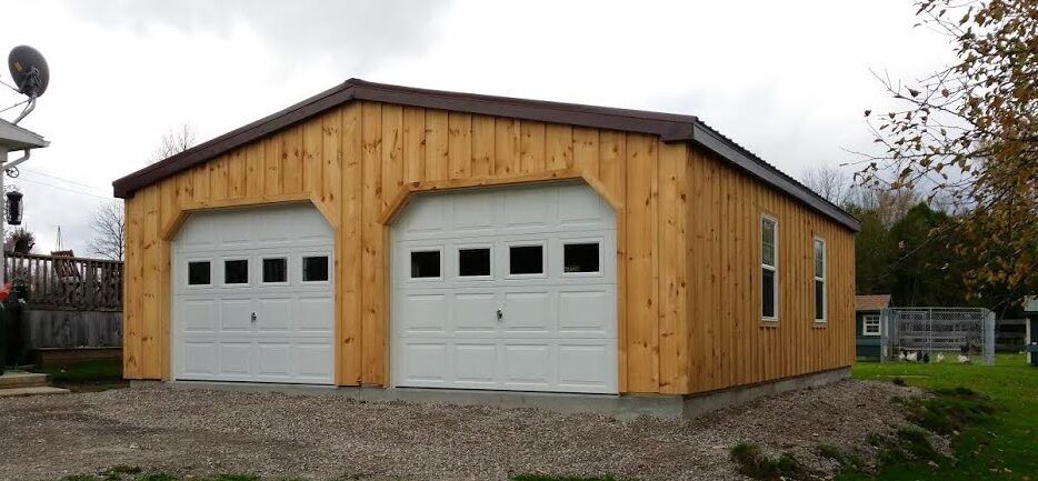 Wooden detached garage with double white doors and pitched roof, built on gravel foundation in backyard.
