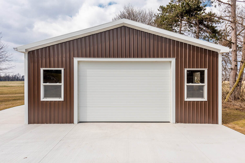 Brown metal garage building with white roll-up door and two side windows, built on concrete driveway.