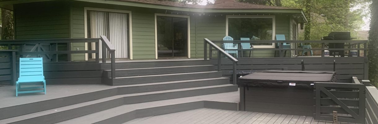 Green cabin with expansive grey deck featuring various outdoor furniture and hot tub